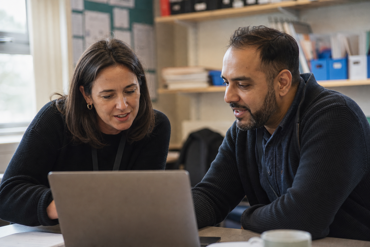 Two teaching colleagues leaning in to look at a laptop together in a staffroom, mid-conversation and at ease.